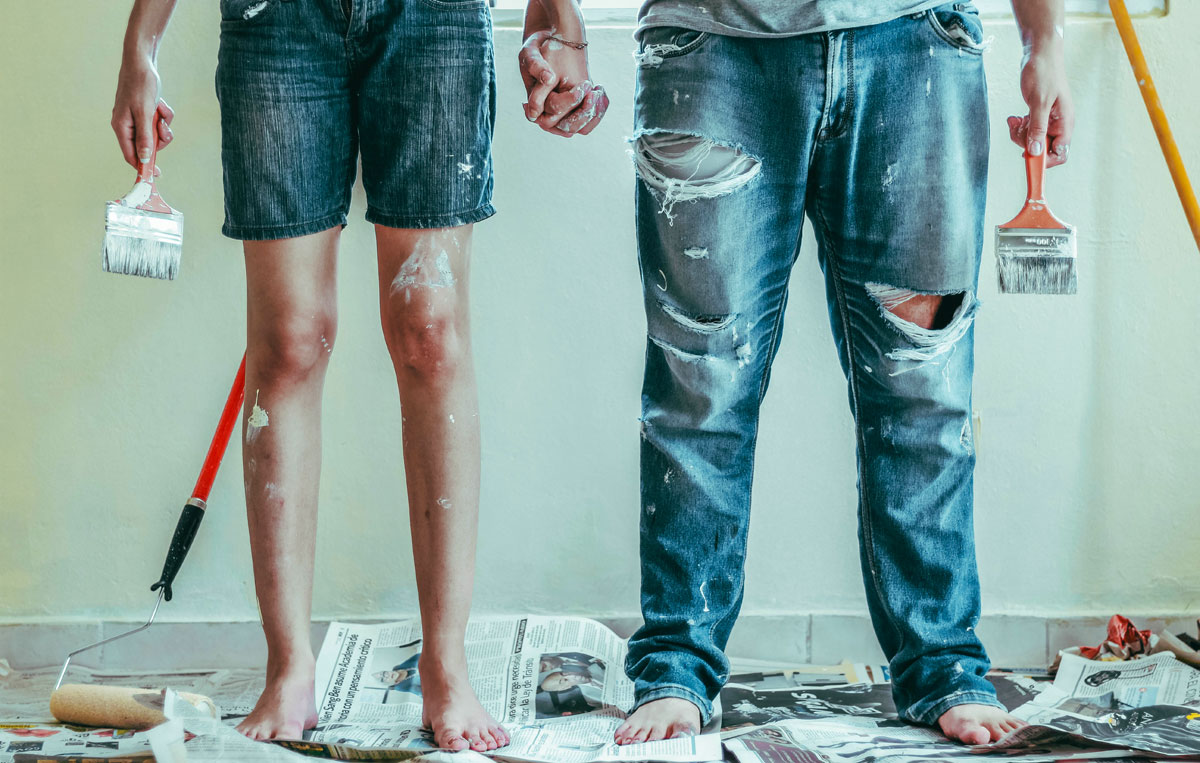 Two people wearing denim holding paint brushes and standing on a pile of newspaper