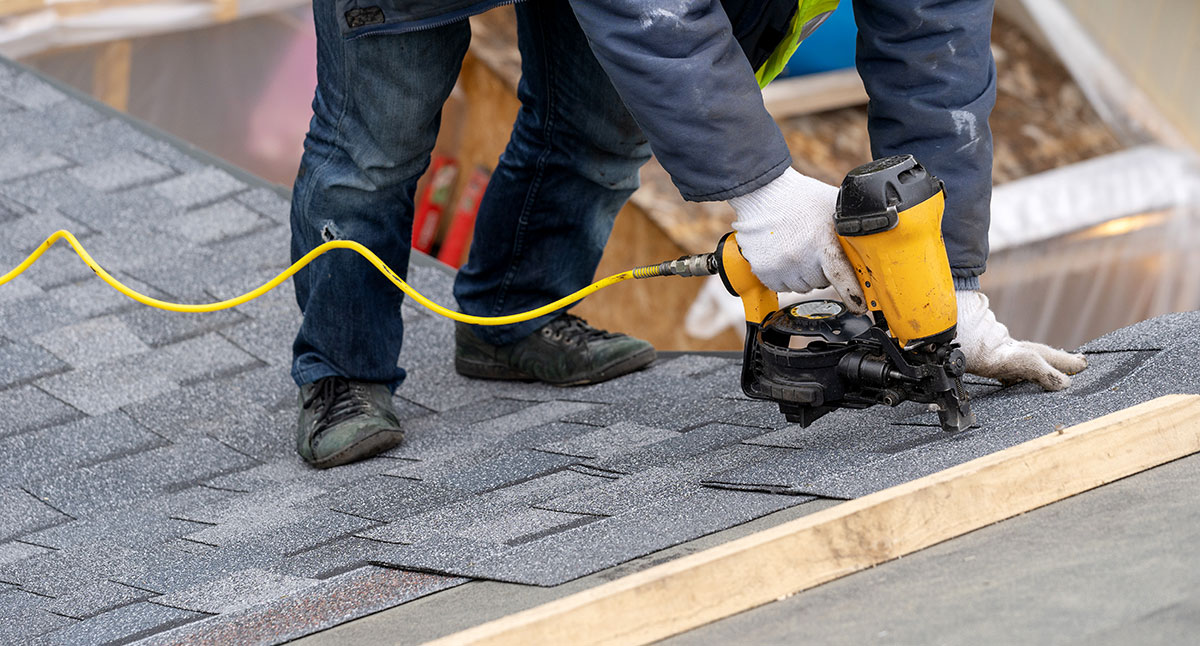 professional roofer worker in uniform work wear using air or pneumatic nail gun and installing asphalt or bitumen shingle on top of the new roof under construction residential building