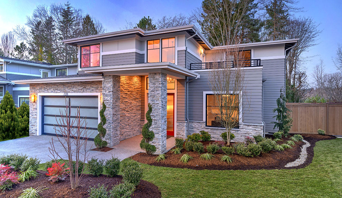 Modern contemporary house exterior with luxury details, landscaping, stone, wood, glass, lots of large windows with evening lights and red front door.