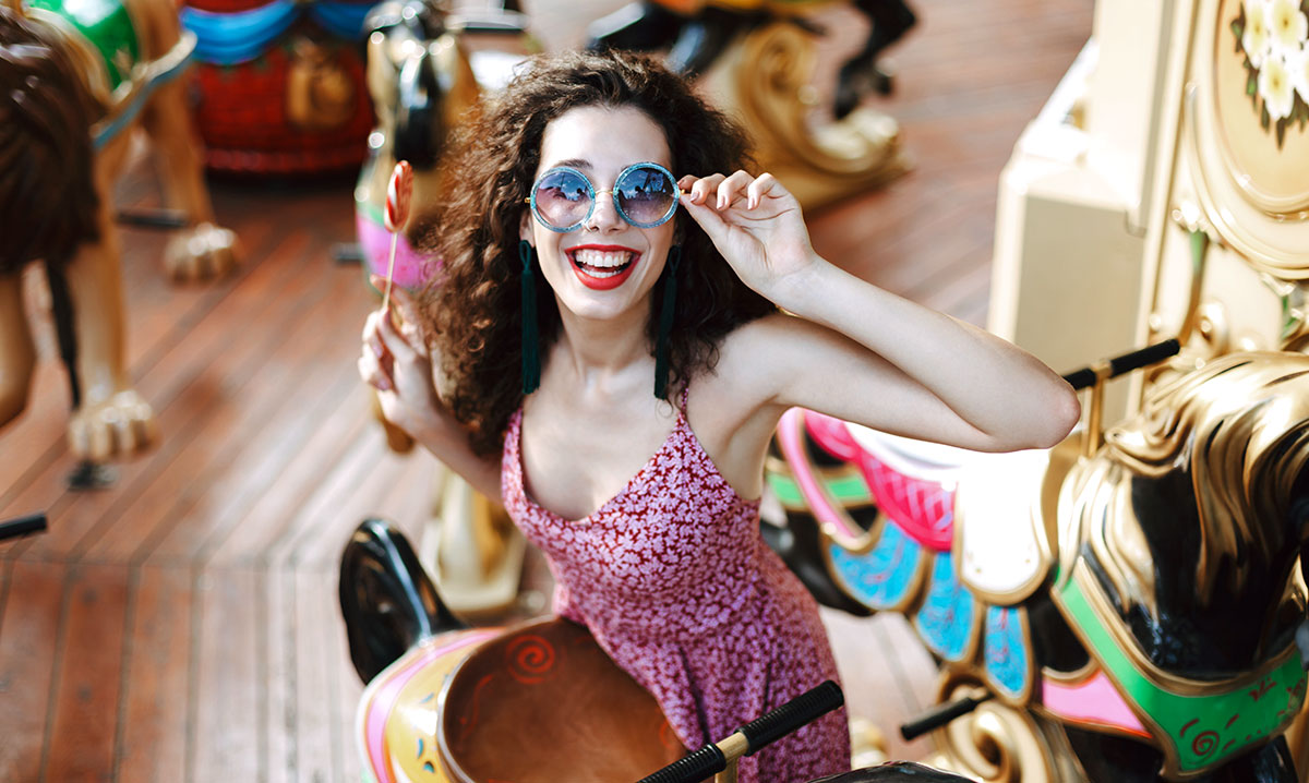 Smiling lady with dark curly hair in sunglasses and dress standing with lolly pop candy in hand and happily looking in camera while riding on carousel in amusement park