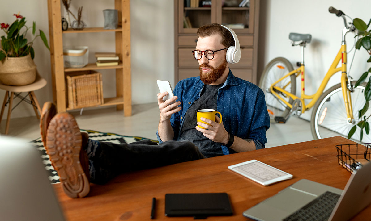 Content bearded designer listening to music in wireless headphones and surfing net while drinking coffee during break in home office