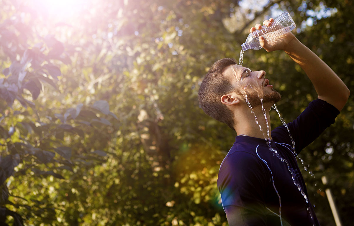 A man pours water over his head to cool off on a sunny day in a leafy outdoor setting.